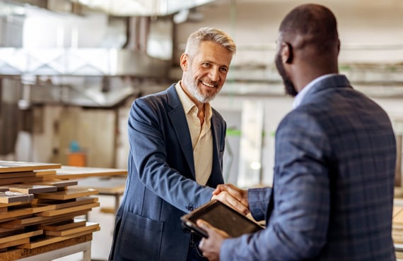 Two business professionals shaking hands in a workshop. Stacks of wooden boards and industrial equipment are visible in the background.