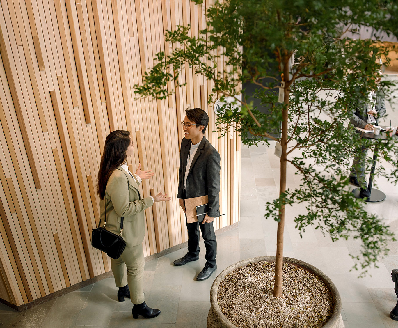 A high-angle view of two professionals engaged in conversation in a modern office lobby, surrounded by wooden panelling and a potted tree.