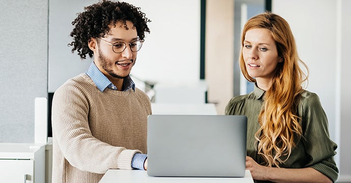 Two colleagues stand at a filing cabinet viewing a laptop screen in a bright office space.
