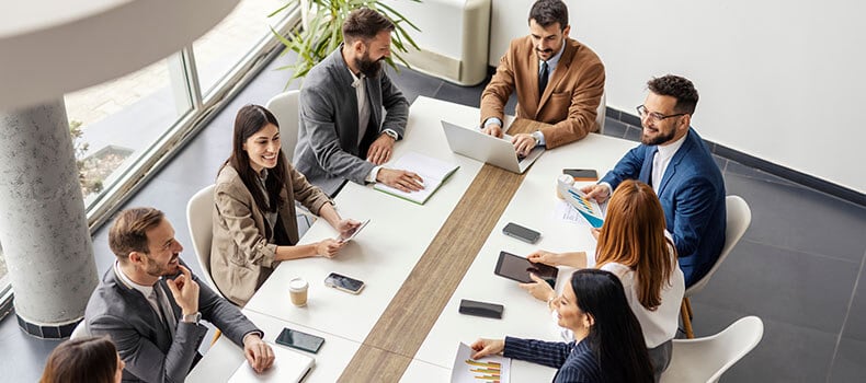Huit professionnels (hommes et femmes) sont assis à une table de réunion avec leurs ordinateurs portables, leurs tablettes, leurs téléphones, leurs documents et leurs graphiques.