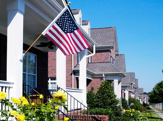 The front porch of a home with an American flag flying and bright yellow roses in the front, with a row of similar homes in the background.