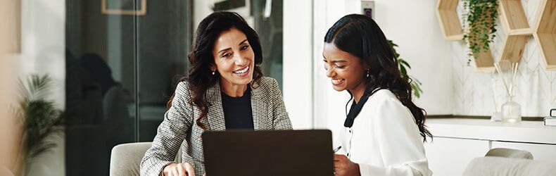 Two business professionals sit at a conference table with a laptop and papers in a bright office with plants and wood shelving.