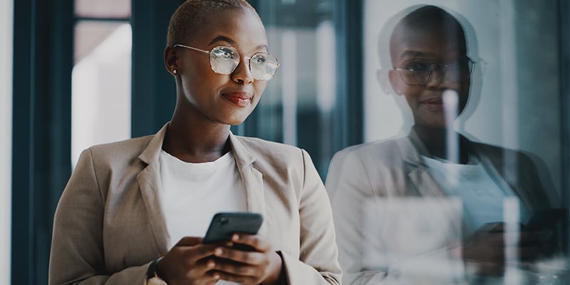 Une femme d’affaires debout devant une fenêtre de bureau utilise un téléphone cellulaire.