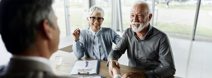 Un couple d’aînés assis à leur table de cuisine serre la main à un associé d’affaires tout en souriant.