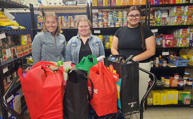 Two BDO staff members smiling behind a cart filled with reusable grocery bags of food, inside a pantry with shelves of canned goods and supplies.