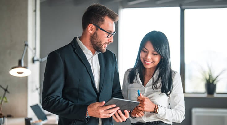 A male and female worker standing next to each other in business attire and looking at a tablet in a modern office environment.