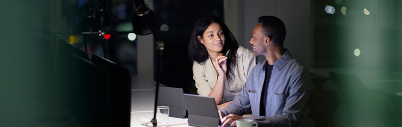 A man and a woman collaborating on laptops in a dimly lit office setting during nighttime.