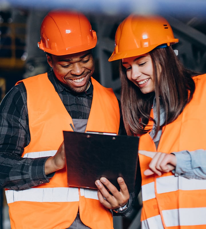 Two construction workers in safety gear holding a clipboard and looking at it in a warehouse setting.