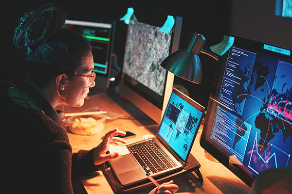 Woman working on a laptop surrounded by three other computer screens depicting information.