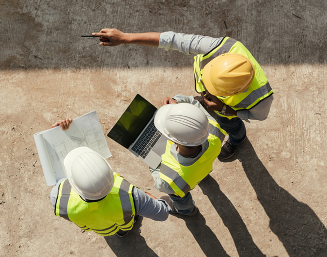 Vue en plongée de trois travailleurs portant des casques de chantier et des vestes de sécurité sur un chantier de construction, le premier tenant des plans, le deuxième tenant un portable et le troisième pointant quelque chose.