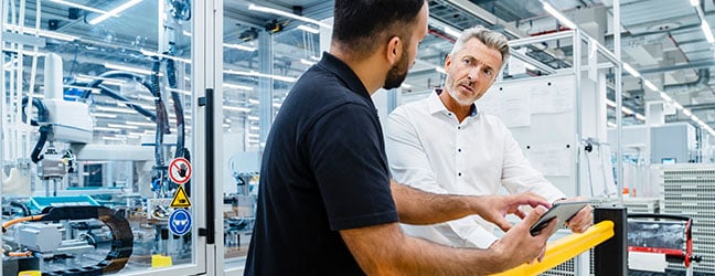 Deux professionnels du secteur de la fabrication discutent dans une usine moderne en tenant une tablette.