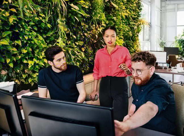 Three employees, a woman standing behind two seated men, looking at a screen in an office setting.