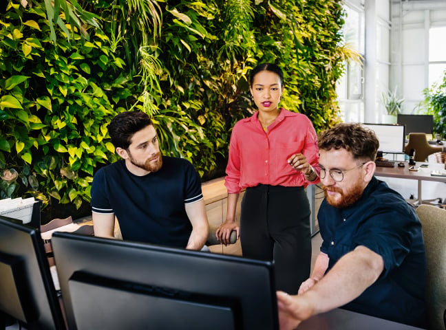 Three employees, a woman standing behind two seated men, looking at a screen in an office setting.