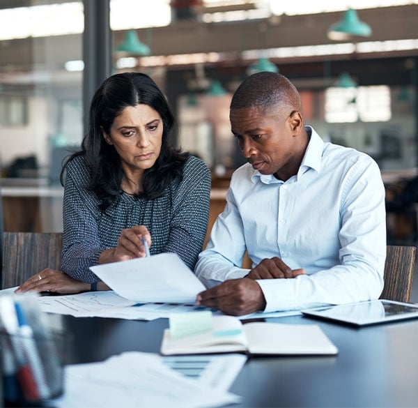 A businesswoman and businessman sitting at a table while looking over paperwork in a modern office.