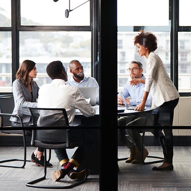 A side view of a female business professional standing in a meeting room addressing a group of four professionals, sitting at a table with open laptops.