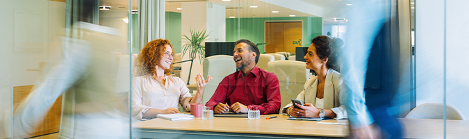 Three employees, two women and one man sitting together and chatting at a table in an office setting.