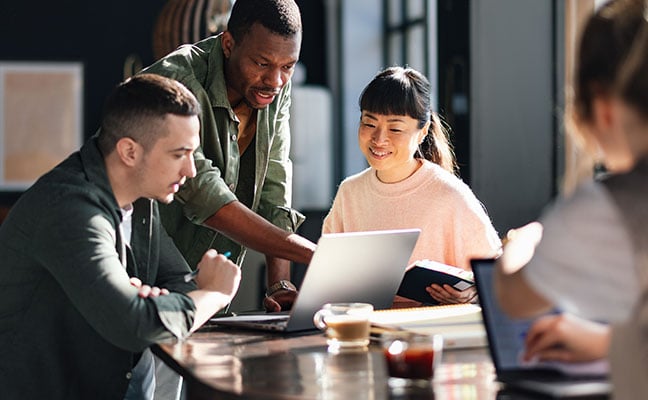 Four coworkers sitting together at a table in an office, with one person pointing at a laptop screen while others take notes and discuss ideas.