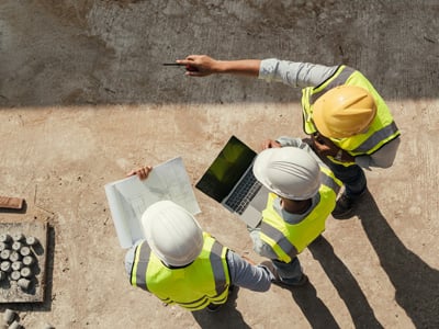 Vue en plongée de trois travailleurs debout sur un chantier portant un gilet de sécurité haute visibilité et un casque de sécurité. L’un des travailleurs tient un ordinateur portable et un autre tient des plans.