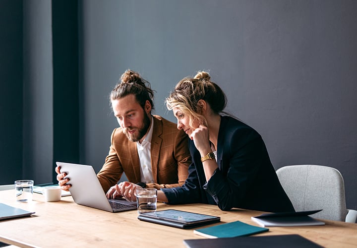 Two young business people, a young man and a young woman, sitting at a desk working on a laptop and a tablet.