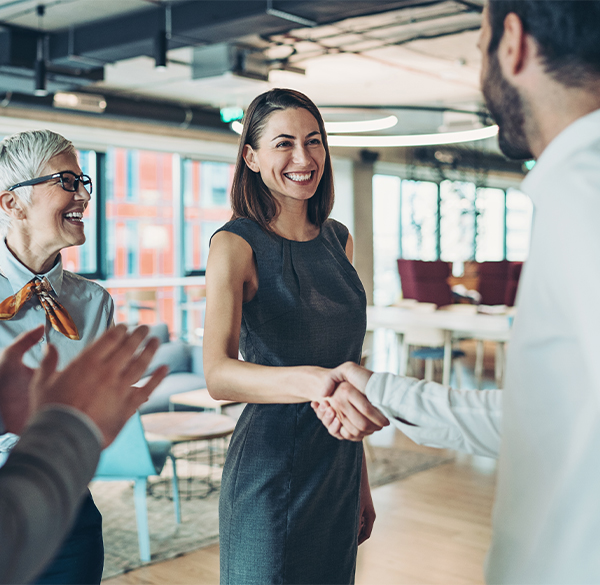 A female and male business professional standing and smiling at each other while shaking hands in a bright office setting. Another female business professional stands next to them and smiles at her male colleague.