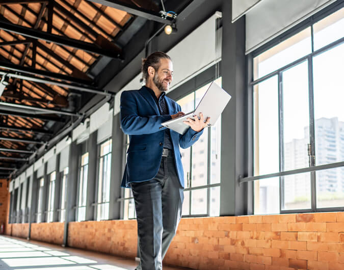 A businessman walks through an empty office while looking at a laptop screen.