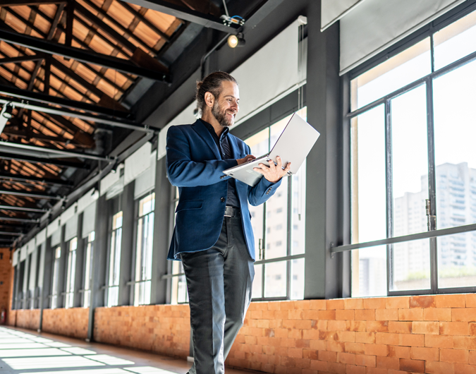 A businessman walks through an empty office while looking at a laptop screen.