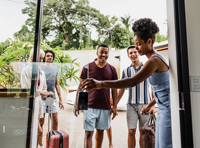 A woman opens a sliding glass door to welcome a group of people with luggage standing outside.