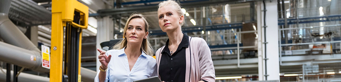 Deux professionnelles, l’une une tablette à la main, discutent en marchant dans une usine.