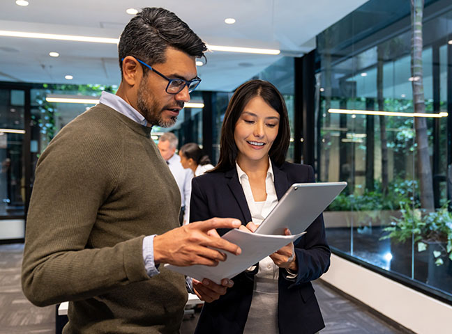 Dans un bureau, un homme et une femme debout regardent des documents et une tablette.