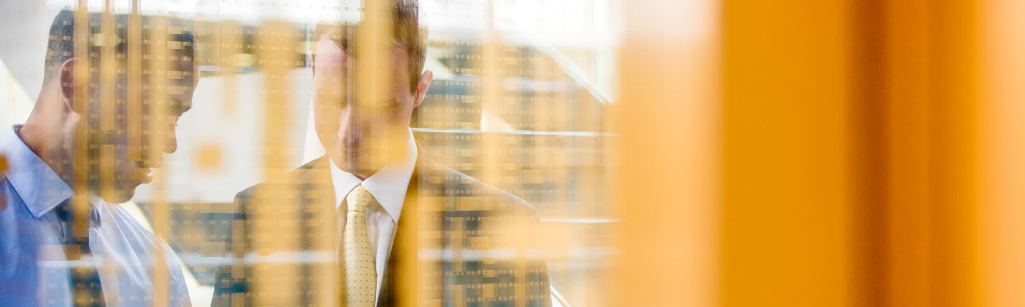 Shot through a glass wall, two men in business attire looking down at a screen.
