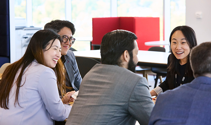 Five employees sitting at a table and engaging in a discussion in a work setting.