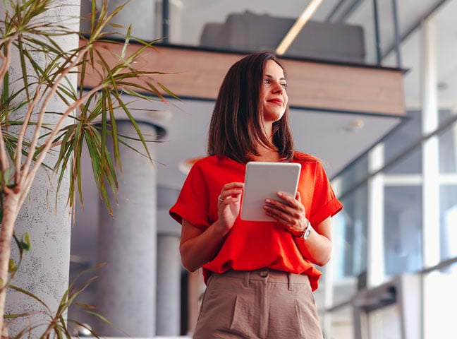 A female professional holds a tablet and smiles in a modern office with a large plant beside her.