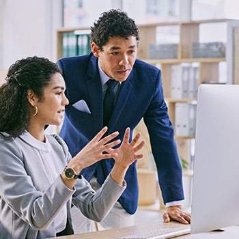 A standing male professional and a seated female professional discussing, looking at a computer, in an office setting.
