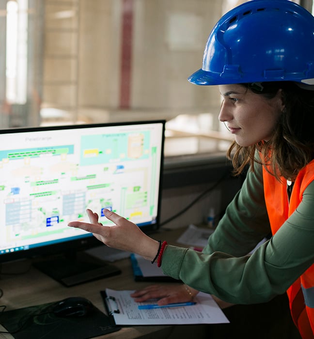 Une ingénieure qui porte un casque de construction et un gilet de haute visibilité regarde deux écrans dans une usine.