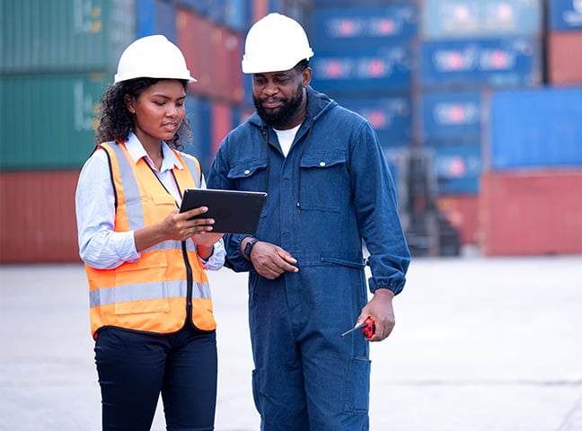 Female port engineer and dock worker are discussing over a tablet computer to organize shipments via mobile app