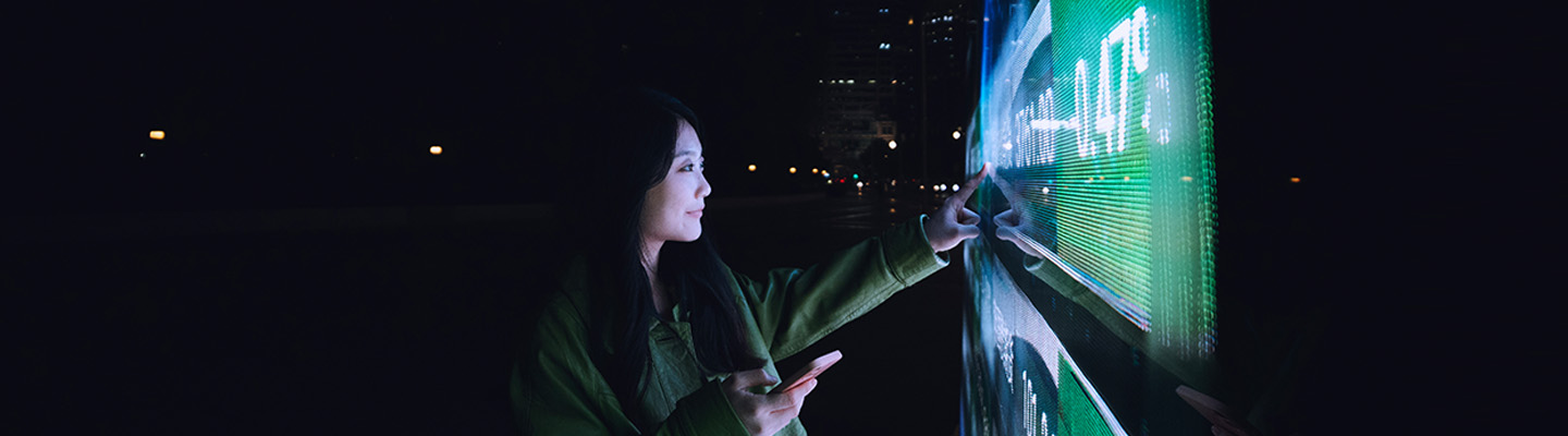 Woman standing outdoors at night interacts with a digital screen.