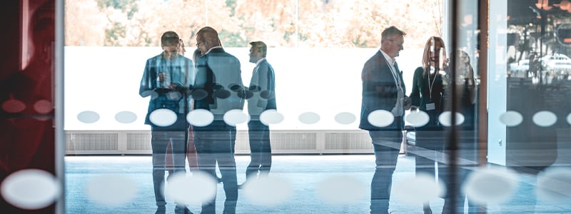 À travers une fenêtre en verre, trois groupes de gens d’affaires debout se parlent.