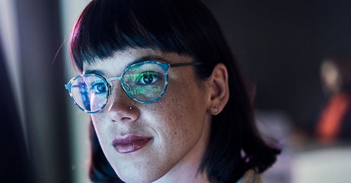 Close-up of a woman's face with data on a screen reflecting in her glasses.