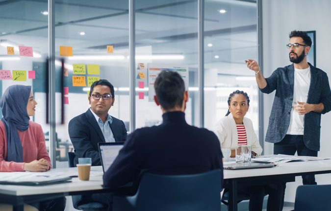 A group of business professionals engaged in a discussion in a modern meeting room. One is standing at the front of the room next to a large monitor with graphs and charts displayed.