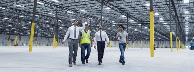 Two business men in suits and two workers in protective gear walking and talking in a large, empty warehouse.