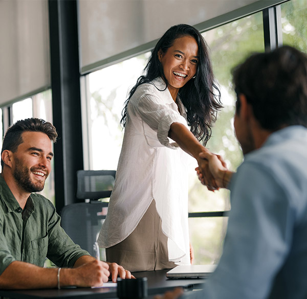 A man and a woman in casual business clothing smiling and facing another man in a modern office environment. The woman is standing up and shaking hands with the man seated across from her. The man next to her is sitting down.