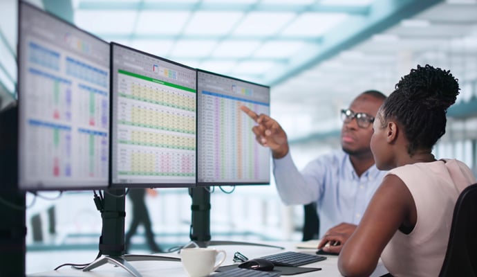 Two man and woman in business attire sit at a desk in an office, viewing charts and graphs across a three-monitor setup.