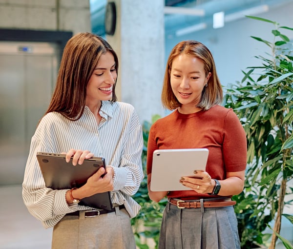 Two women standing in a modern office space smiling and both looking at a tablet. The space has an industrial design with exposed concrete.