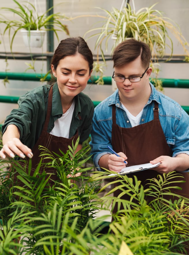 personne trisomique écrivant des notes tout en aidant un collègue avec des fleurs en serre