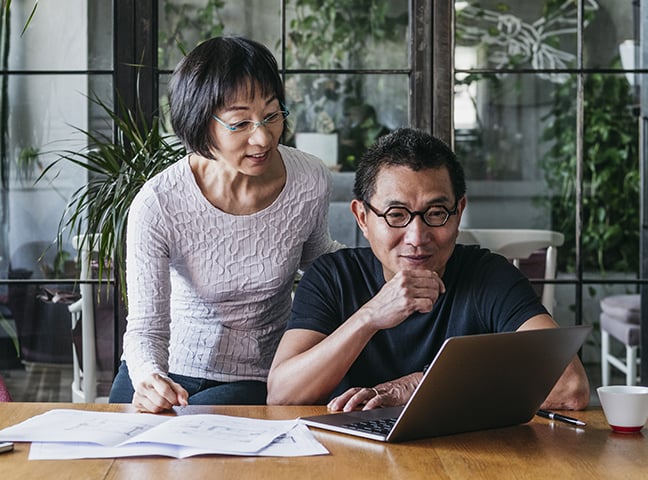 An older couple both looking at a laptop on a wooden table. The man is sitting down while the woman is standing next to him and leaning forward. There are a few documents beside the laptop. Large windows and plants are in the background.