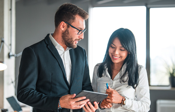 A male and female worker standing next to each other in business attire and looking at a tablet in a modern office environment.