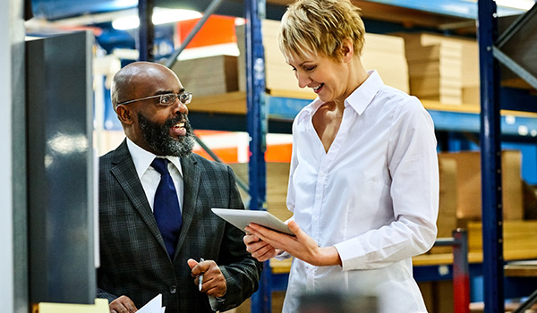 Female and male professionals are talking, one holding a tablet, in a warehouse setting, depicting smart manufacturing context.
