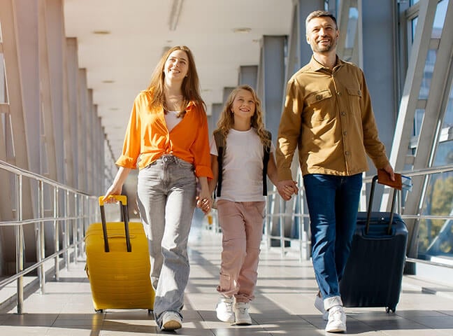 A smiling family of three walks through a bright airport corridor with large windows. Both parents are each pulling a rolling suitcase and holding the hands of a young girl between them.