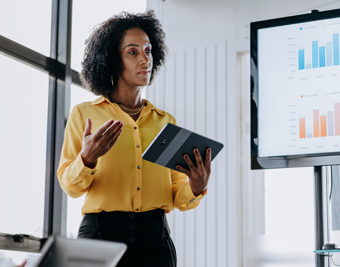 A businesswoman holding a tablet stands next to a large monitor with charts displayed in a modern meeting room.