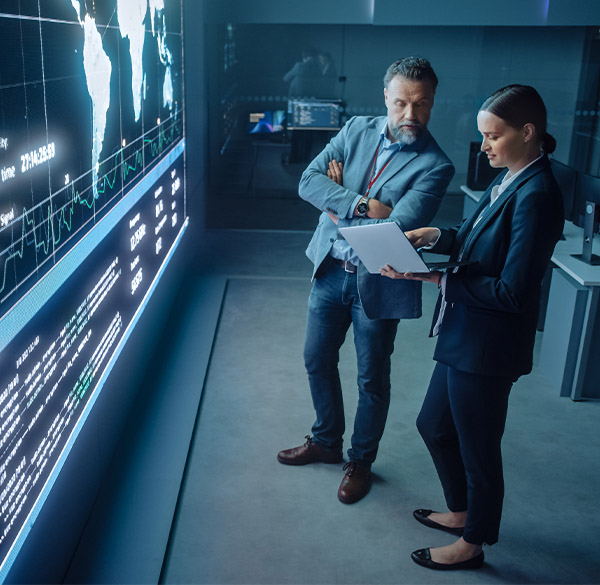 A man and a woman standing and both dressed in business attire while looking at a laptop. They’re both in front of a large screen with a map of the world displayed on it in a darkened room.